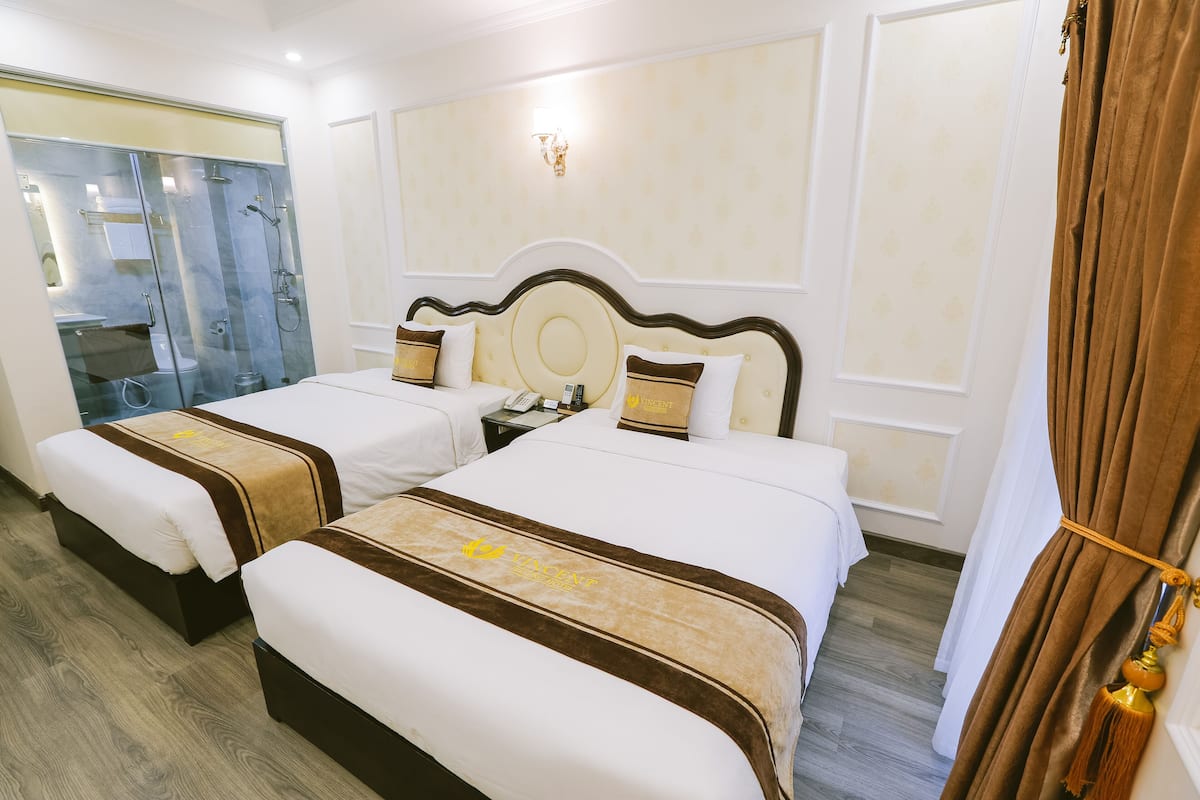 A hotel room with two beds, a bathroom with a glass shower enclosure, and a large window with curtains.