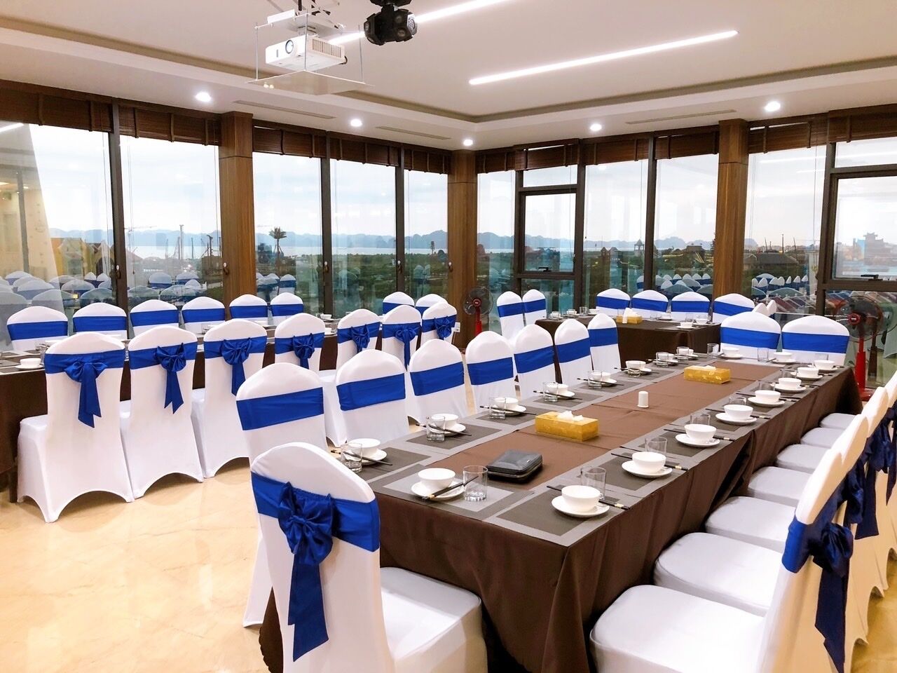 A conference room with a long table set for a meeting, surrounded by chairs with blue sashes, large windows with a city view, and a modern ceiling design.