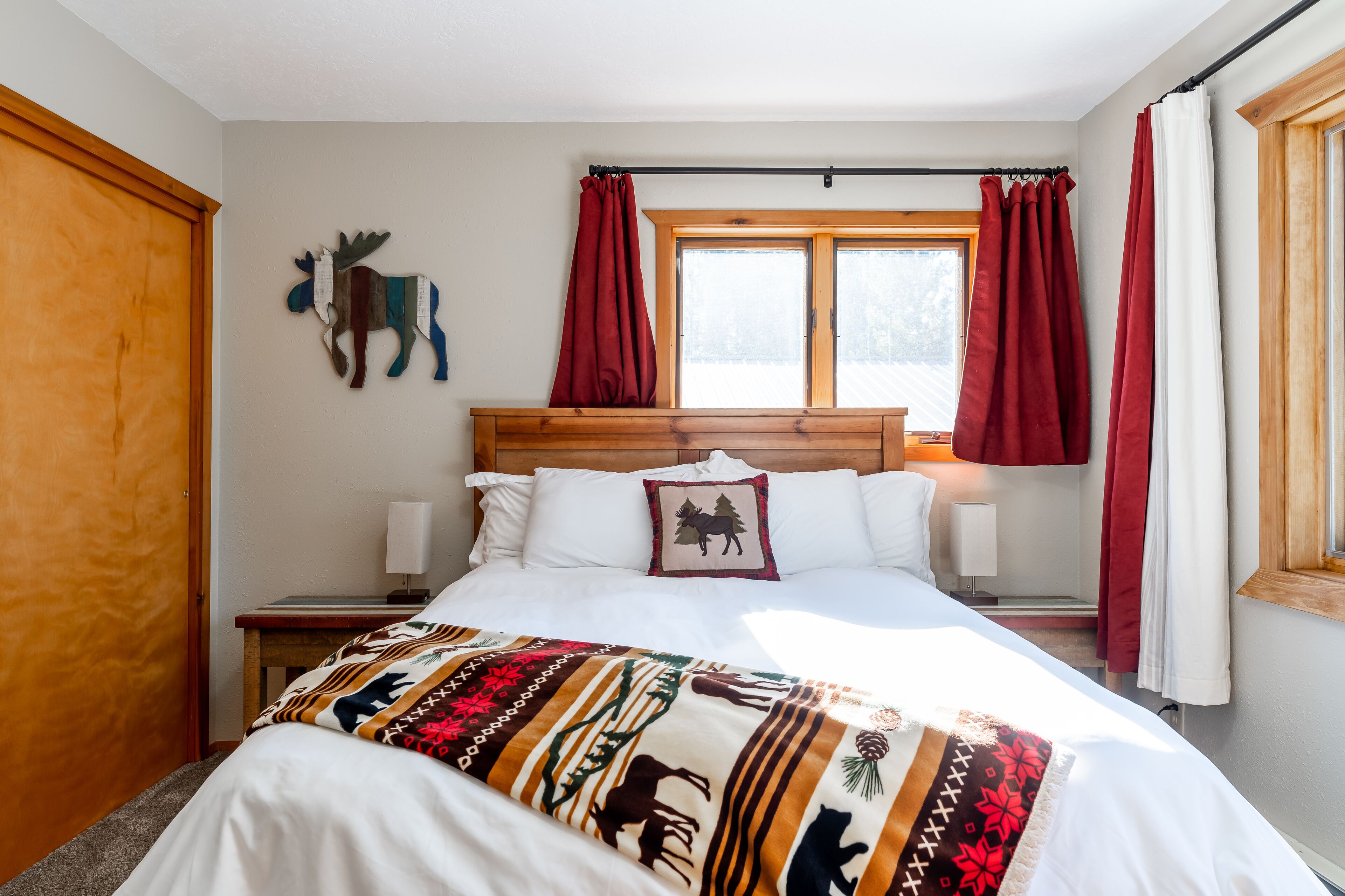 A bedroom with a wooden headboard, a bed with a patterned blanket, two bedside lamps, a window with red curtains, and a wall-mounted moose decoration.
