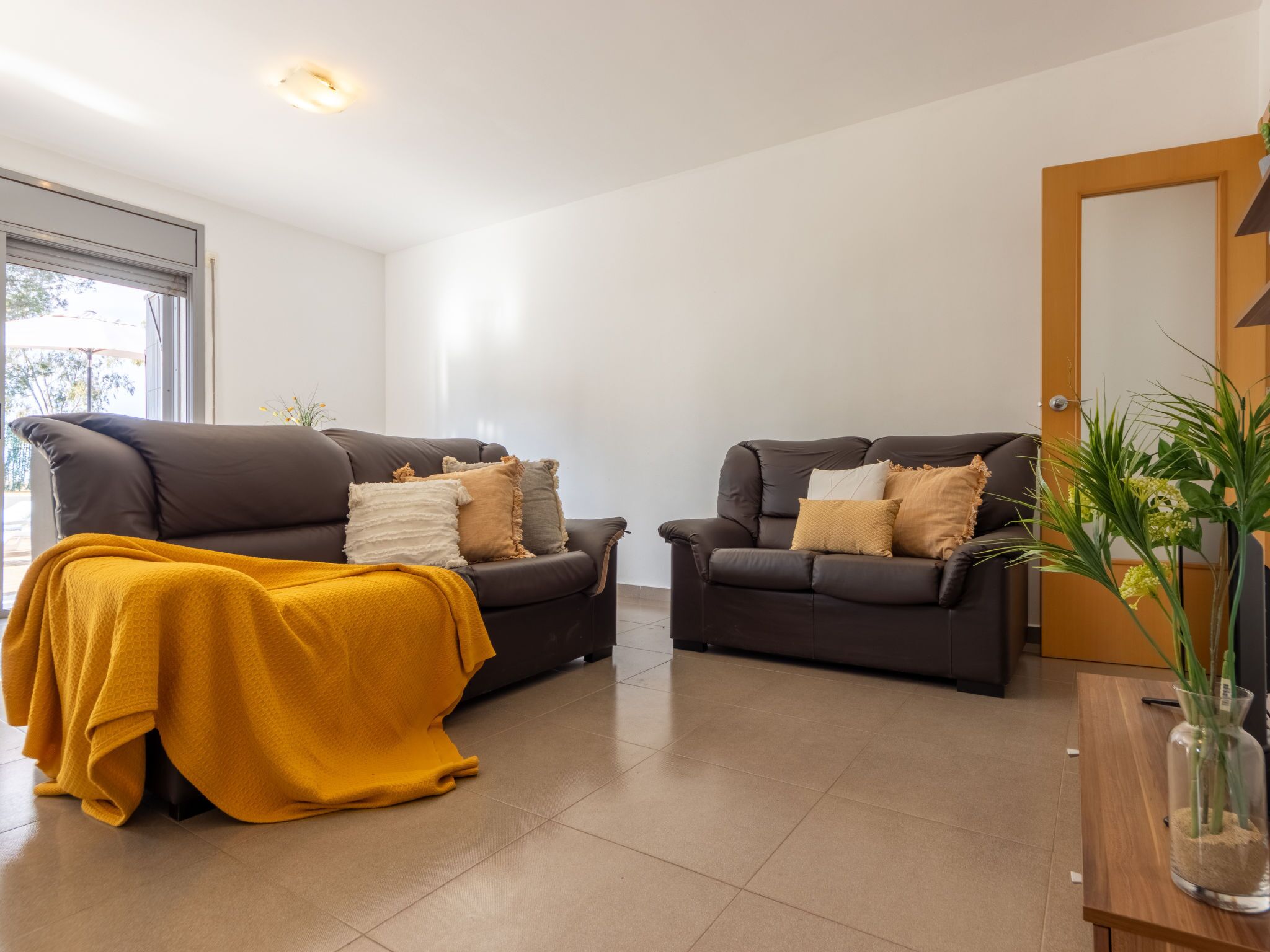 Una sala de estar moderna con dos sofás marrón oscuro, una manta amarilla y una mesa de centro de madera con un jarrón de plantas verdes.