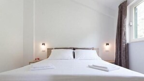 Una cama bien tendida en una habitación de hotel con sábanas y almohadones blancos, dos mesitas de noche con lámparas y una ventana con cortinas.
