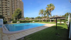 A swimming pool with a tiled deck, surrounded by well-maintained grass and palm trees. - Apto CasaTuris 1Línea Muchavista C101 (Ibi)