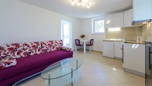 Una sala moderna con un sofá púrpura, una mesa de centro de vidrio, un comedor con una mesa pequeña y sillas, y una cocina con armarios blancos y una estufa.