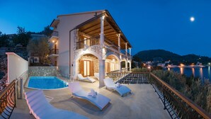 A two-story villa with a swimming pool, lounge chairs, and a view of the lake at dusk. - Villa Amfora- Six Bedroom Villa with Terrace and Swimming Pool, 2X Jacuzzi and Sea View (Prižba)