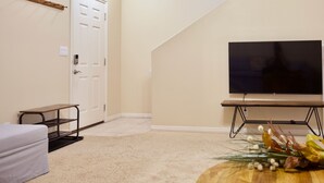 Una habitación con una televisión de pantalla plana sobre un soporte, una puerta, un pequeño banco y una planta decorativa.