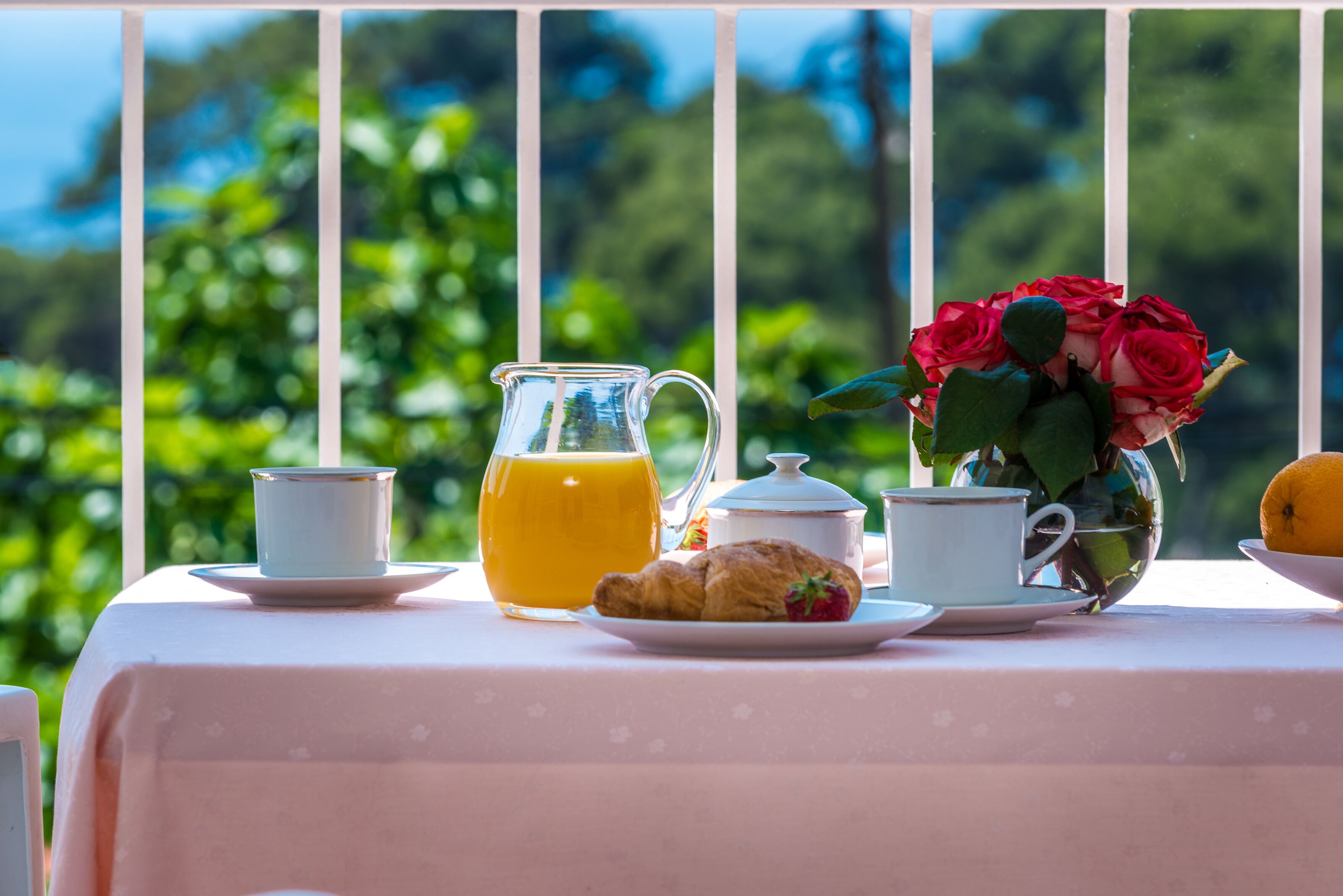 A breakfast table with a pitcher of orange juice, a croissant, a teapot, and a vase of red roses.
