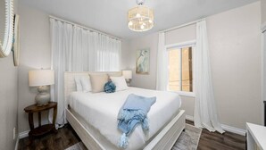 Ein Schlafzimmer mit einem großen Bett, einem Nachttisch, einer Lampe, einem Fenster mit Vorhängen und einem Wandbild.