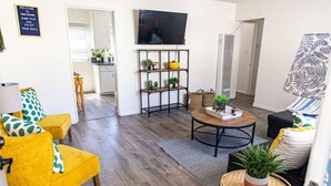 A living room with a yellow armchair, a round coffee table, a TV mounted on the wall, and a bookshelf.