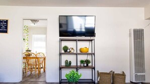 A living room with a TV on a black metal stand, a dining area with a wooden table and chairs, and a white air conditioning unit.