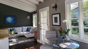 A modern living room with a glass dining table, a sofa, and a large window with a view of greenery. - Private Cottage Oasis in CdM - Ideal Village Location on XL Lot (Corona del Mar)
