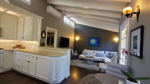 A modern kitchen with white cabinetry, a built-in microwave, and a large island. There is a dining area with a white sofa and a coffee table. The room has a dark wall with a blue painting and a framed picture. - Private Cottage Oasis in CdM - Ideal Village Location on XL Lot (Corona del Mar)