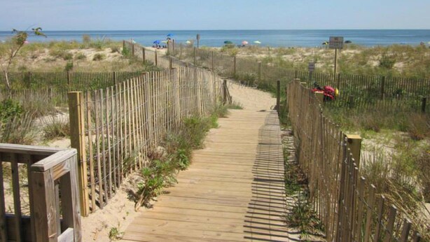 Un muelle de madera que conduce a una playa, con una valla a un lado y un sendero de arena al otro.