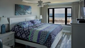 Un dormitorio con cama, mesita de noche, escritorio, ventilador de techo, ventana con vista a la playa y un cuadro en la pared.