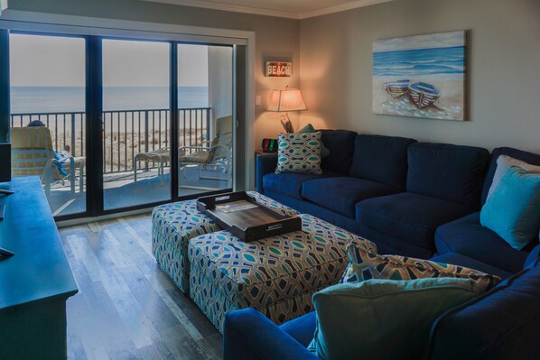 Un salón con un sofá azul, un reposapiés estampado y vistas a la playa a través de un ventanal corredero.