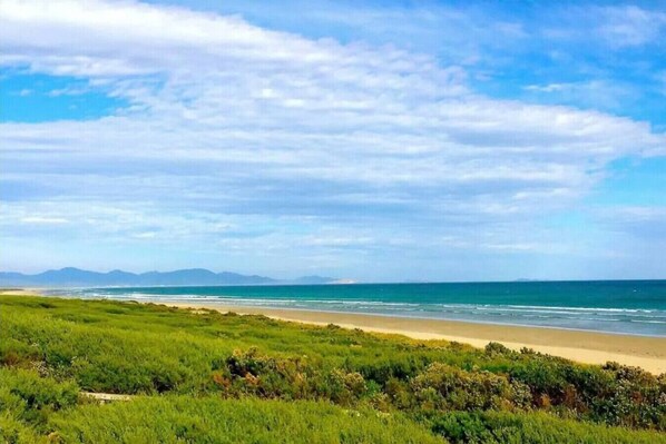 A coastal landscape with a beach, grassy foreground, and distant mountains under a partly cloudy sky.