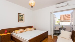 Une chambre avec un lit en bois, une table de chevet avec une lampe rouge, un tableau encadré au mur et un balcon donnant sur des immeubles.