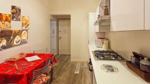 A narrow kitchen with a gas stove, a microwave, and a kettle. There is a dining table with a red tablecloth and a chair. The walls are adorned with artwork depicting food and drink. - Venice Dream House Figaro (Venice)