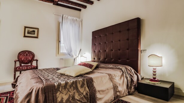 A bedroom with a large bed, a wooden headboard, a bedside table with a lamp, a chair, and a window with curtains. - Venice Dream House Figaro (Venice)