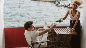A couple seated at a wooden table on a balcony overlooking a body of water with boats and mountains in the background. - Seafront Cave Suite (Santorini)