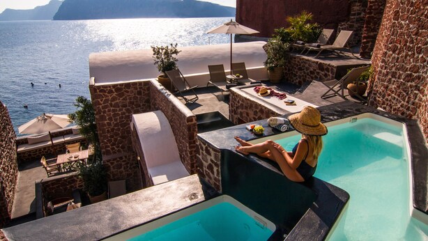 A poolside area with a view of the sea, lounge chairs, and a dining table with umbrellas. - Seafront Cave Suite (Santorini)