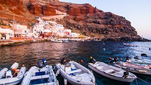 A coastal village with boats docked at the harbor, buildings with white and blue facades, and a rocky cliff in the background. - Seafront Cave Suite (Santorini)
