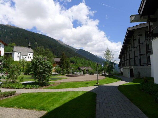 A courtyard with a paved walkway, surrounded by buildings and greenery, with mountains in the background.