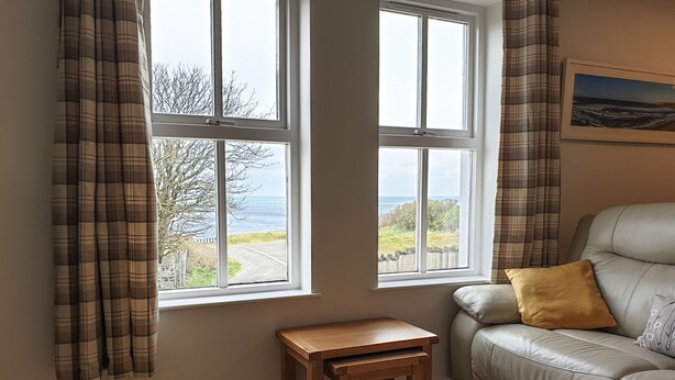 A room with a view of the sea through two windows, a wooden table, and a beige sofa with a pillow. - Coastal View (Bushmills)