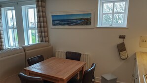 A cozy dining area with a wooden table and chairs, a radiator, a window with a view of the sea, and a small gray trash bin. - Coastal View (Bushmills)