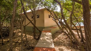 Un petit bâtiment d’un seul étage, avec un chemin en béton menant à l’entrée, entouré d’arbres et avec une plage en arrière-plan.