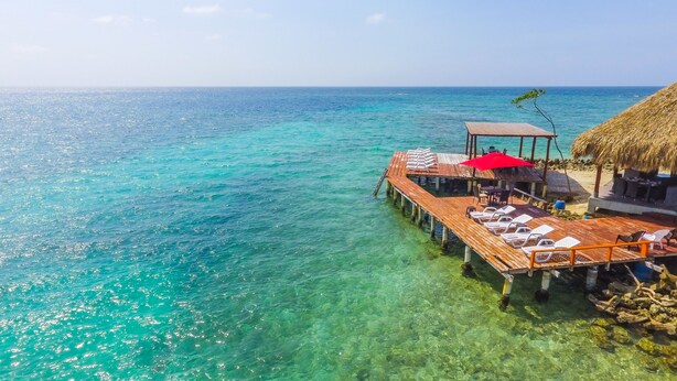 Une jetée en bois s’étendant dans des eaux turquoise et limpides, avec des chaises longues et des parasols sous un toit de chaume.