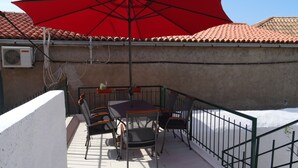 Eine Terrasse mit Tisch und Stühlen, einem roten Sonnenschirm und Blick auf ein Ziegeldach.