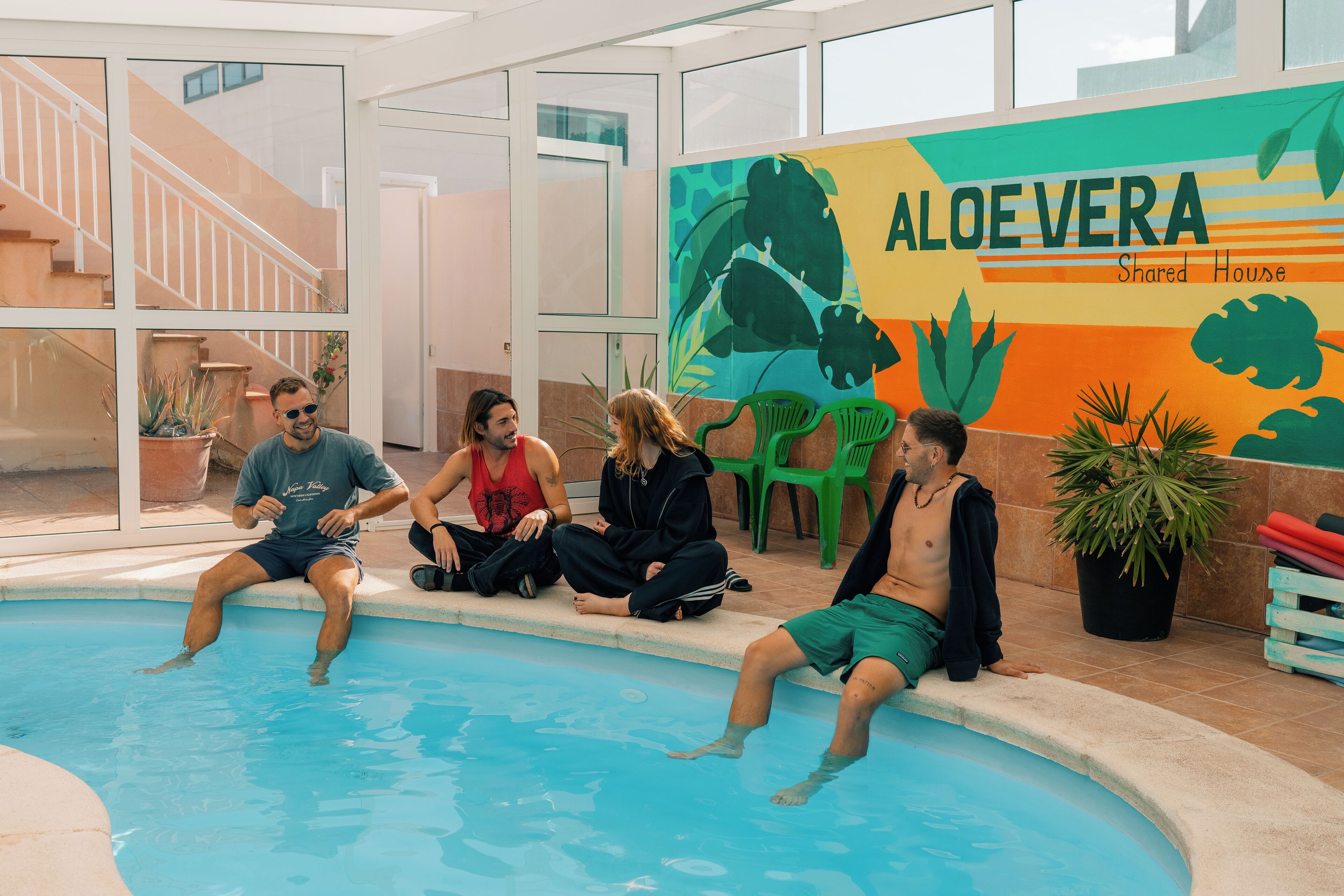 Et poolområde med fire personer siddende ved poolkanten, et mural med teksten 'ALOEE VERA Shared House', og trapper indendørs i baggrunden.