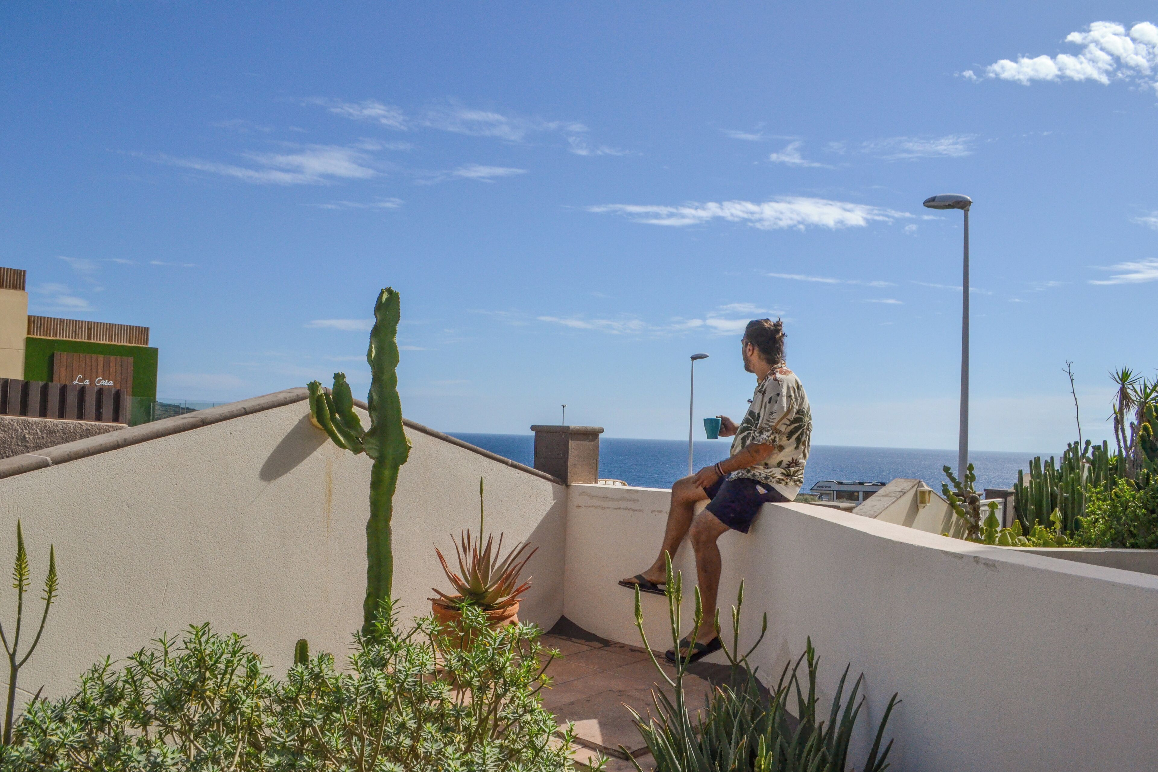 Ein Mann sitzt auf einem Balkon mit Blick auf das Meer, umgeben von Kakteen und Topfpflanzen.