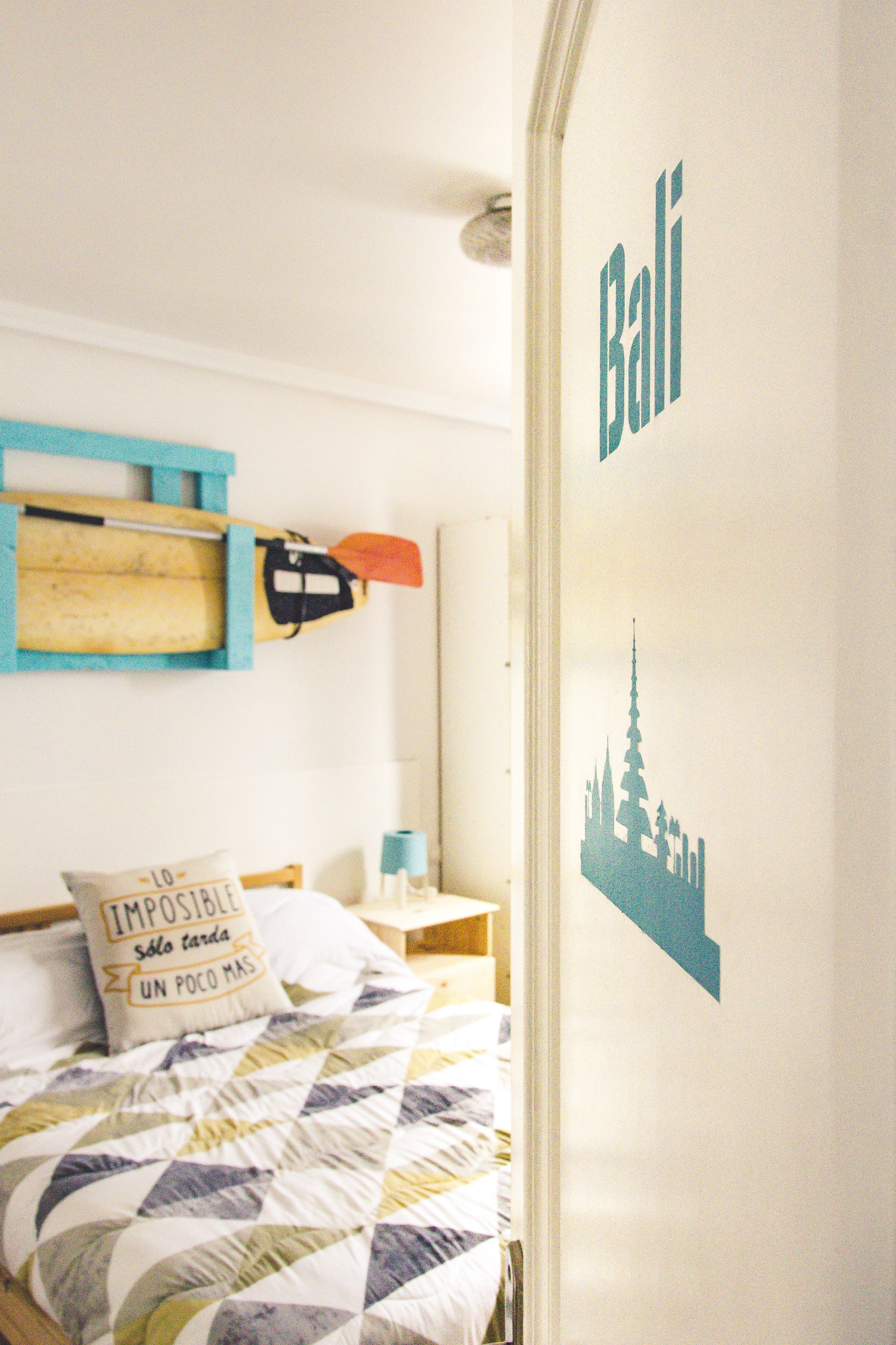 A neatly made bed with a patterned quilt, a pillow with text, a wooden shelf with a paddle, and a wall with a cityscape decal and the word "Bali".