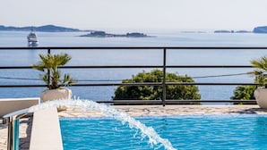 A pool with water cascading over the edge, a view of the sea with a yacht in the distance, and a railing with potted plants.
