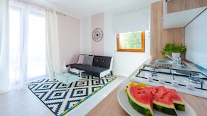 A modern living room with a sofa, a glass coffee table, a patterned rug, a kitchen area with a stove, and a dining table with watermelon slices.