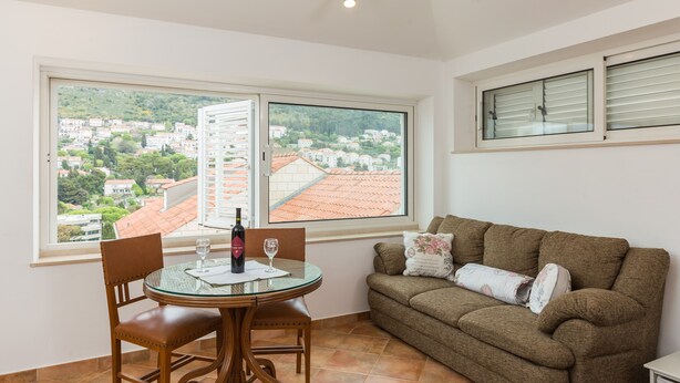 A room with a dining table, a sofa, and a large window offering a view of a town and hills. - Apartments Dia - Studio apartment (Dubrovnik)