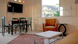 Un comedor con una mesa y sillas, un televisor montado en la pared y una ventana con vistas a una colina.