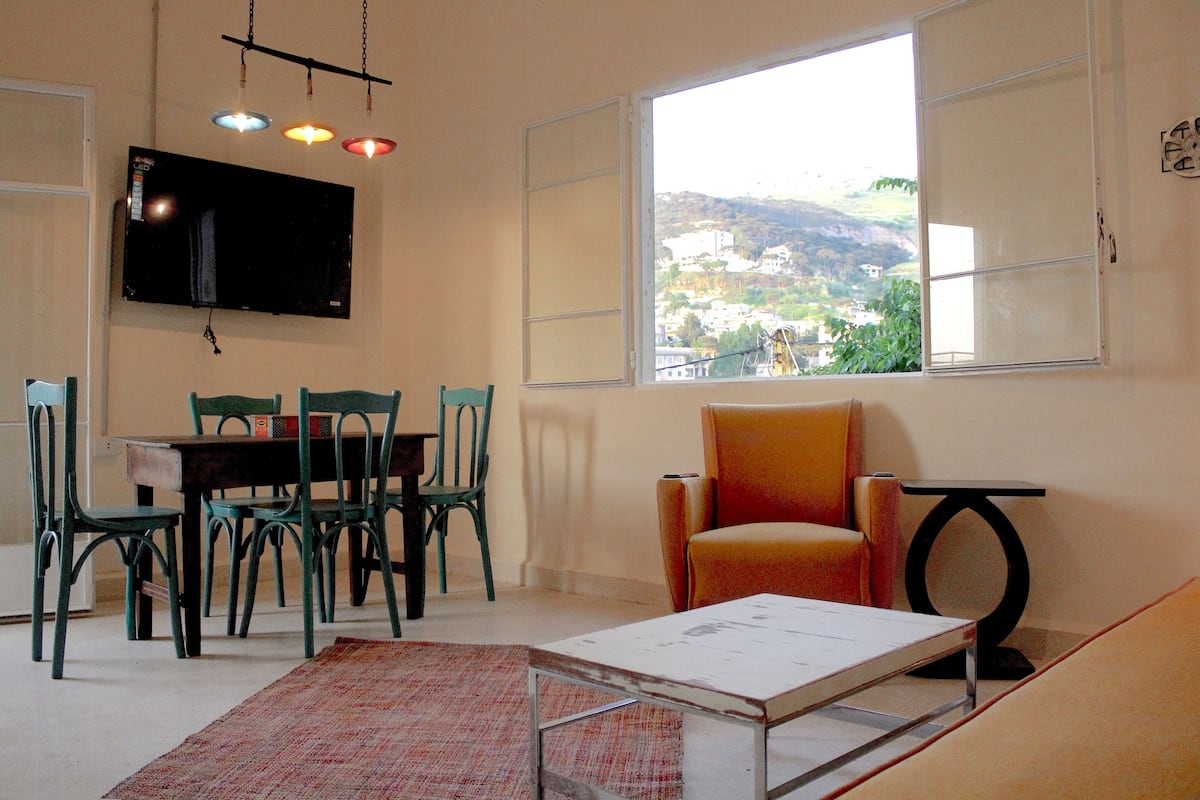A dining area with a table and chairs, a television mounted on the wall, and a window with a view of a hillside.