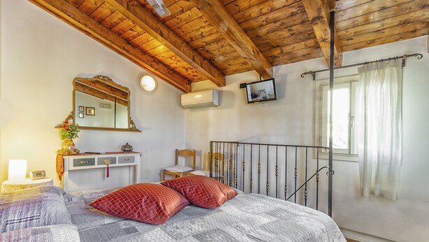 Una camera da letto con soffitto in legno, un letto con biancheria a motivi, un comodino con lampada, uno specchio e un televisore a parete.