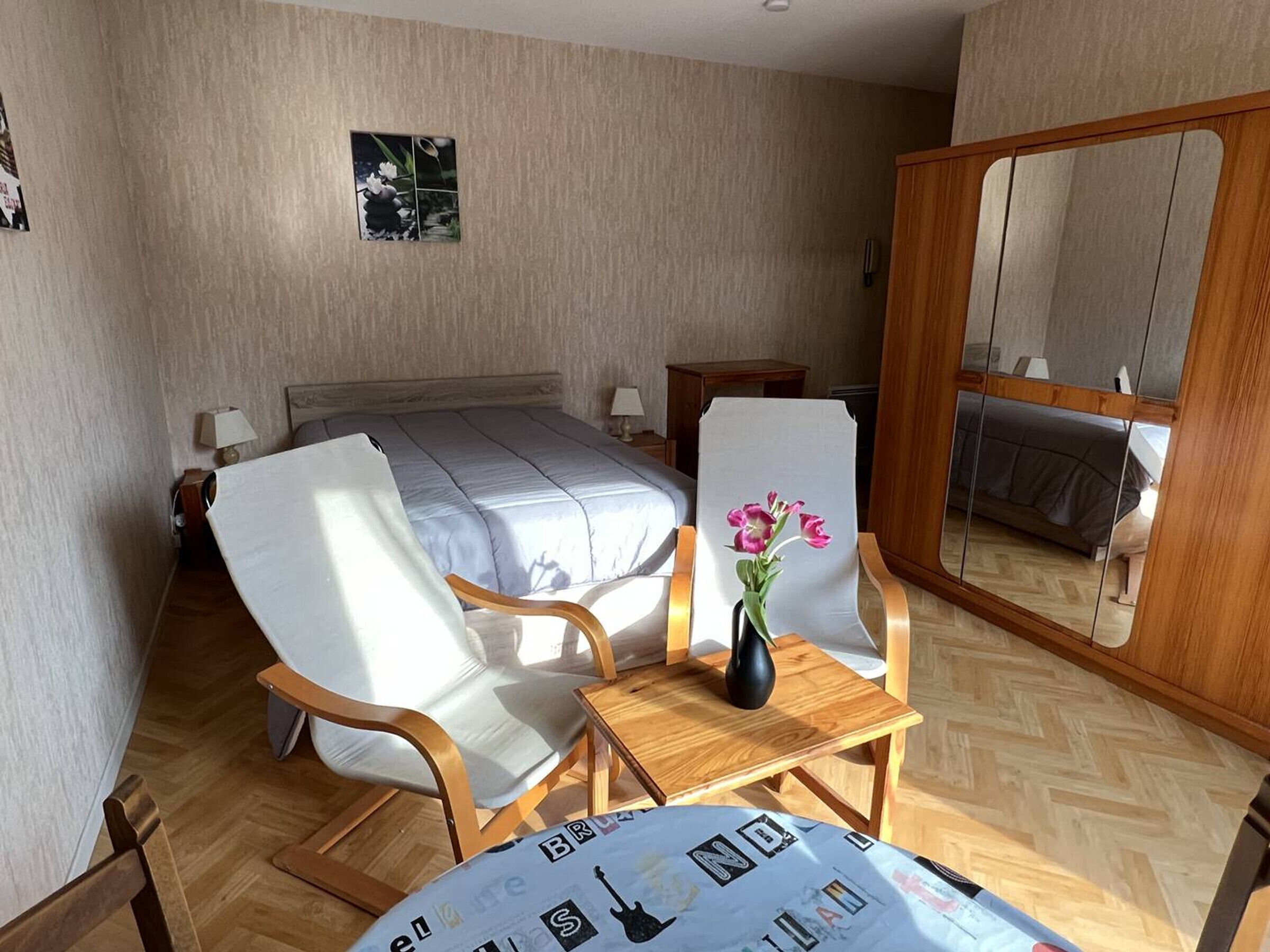 A hotel room with a bed, two chairs, a small table with a vase, and a mirror.