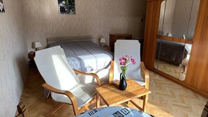 Un dormitorio con cama, dos sillones, una mesita con un florero, un espejo y un cuadro en la pared.