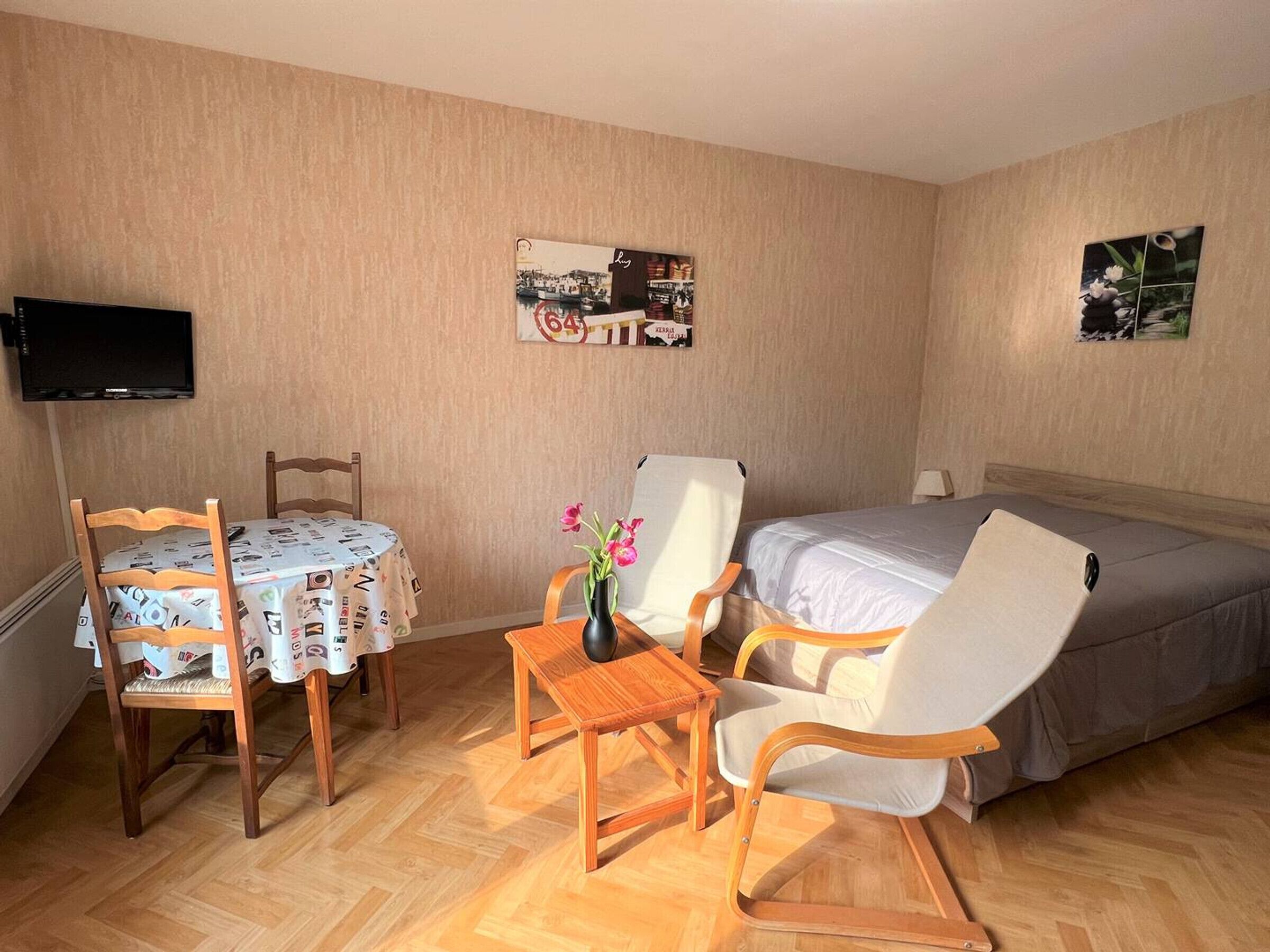 Una habitación de hotel con cama, televisor montado en la pared, mesa de comedor con sillas y una pequeña mesa con un florero.