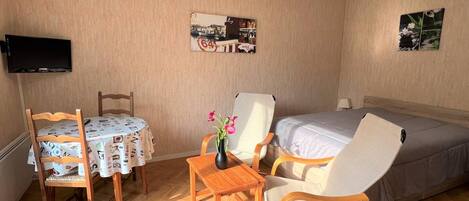 Una habitación de hotel con cama, televisor montado en la pared, mesa de comedor con sillas y una pequeña mesa con un florero.
