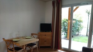 A room with a wooden dining table, two chairs, a television on a wooden cabinet, and a sliding glass door leading to a garden. - Residence Val de Roland - Studio Apartment 2 persons (Luz-Saint-Sauveur)