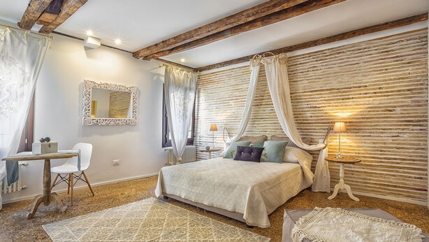 Une chambre avec un grand lit, un bureau et une chaise. Il y a une fenêtre avec des rideaux et une lampe sur une table de chevet.