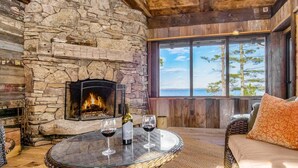 Une pièce chaleureuse avec une cheminée en pierre, une table en verre ornée de verres à vin et d’une bouteille, et une vue sur l’extérieur grâce à de grandes fenêtres.
