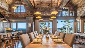 Une grande table à manger en bois, dressée pour un repas, entourée de plusieurs chaises, dans un intérieur rustique avec des poutres apparentes et de grandes fenêtres donnant sur un plan d’eau.