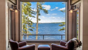 Une pièce dotée d’une grande fenêtre donnant sur un lac et des arbres. On y trouve deux fauteuils en cuir et un petit pouf.