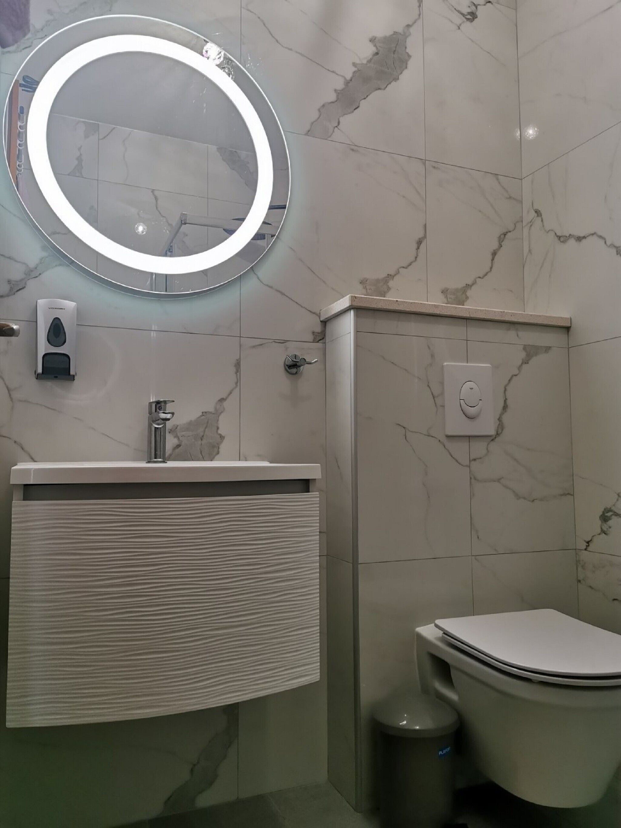 Un baño con pared de azulejos de mármol, un espejo redondo iluminado y un lavamanos moderno, además de un inodoro.